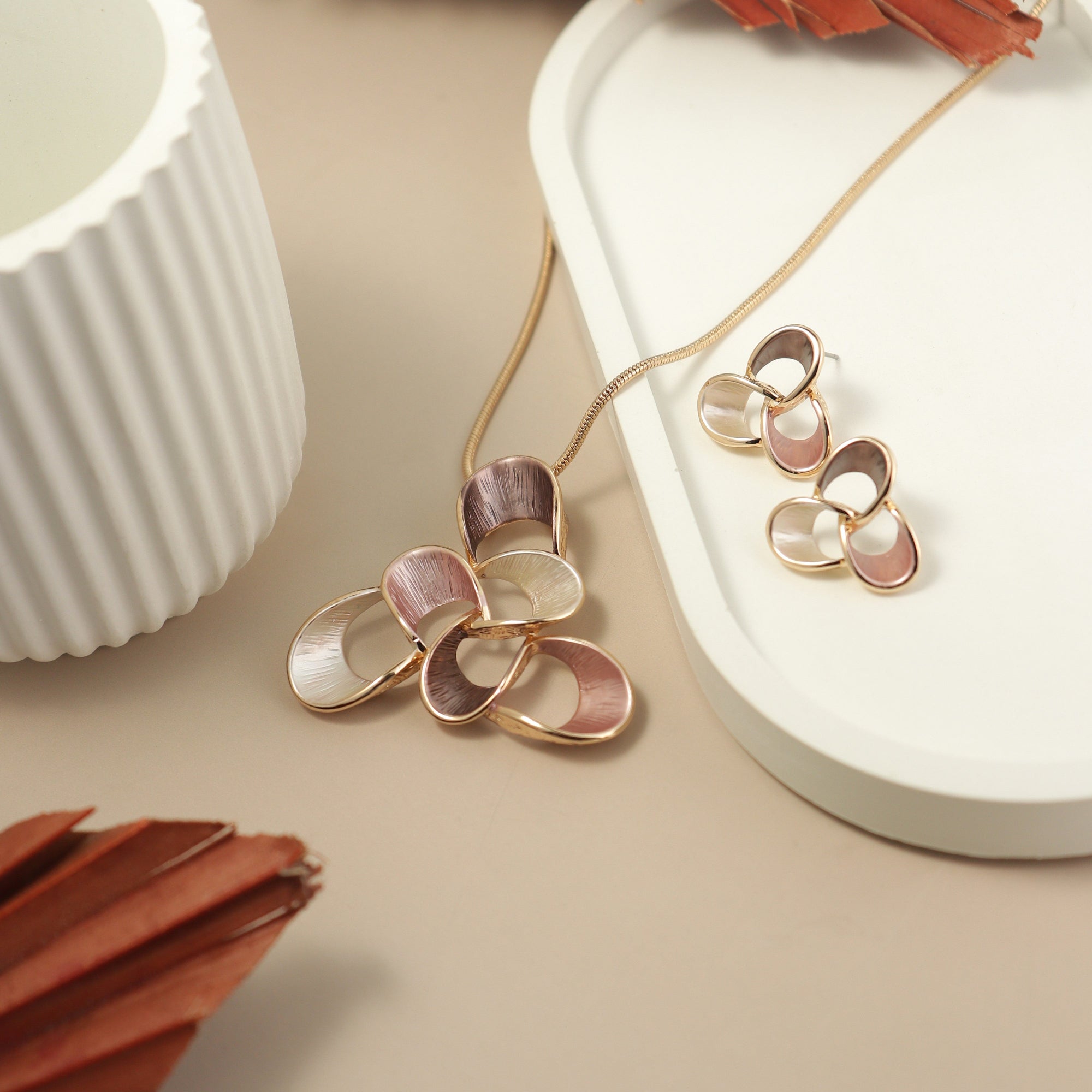 Elegant floral jewelry set featuring a multi-petal necklace and matching earrings in rose gold, blush pink, cream, and mauve tones, displayed on a minimal beige and white background with modern decor elements.