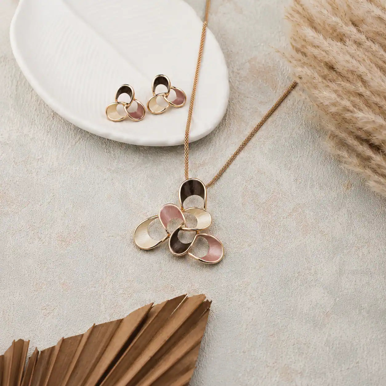 A gold-tone necklace with a flower-shaped pendant featuring alternating metallic gold and pale pink petal-like sections, displayed on a textured light gray surface with a white dish, dried grass, and a folded brown paper fan as props. jewelry for daily use