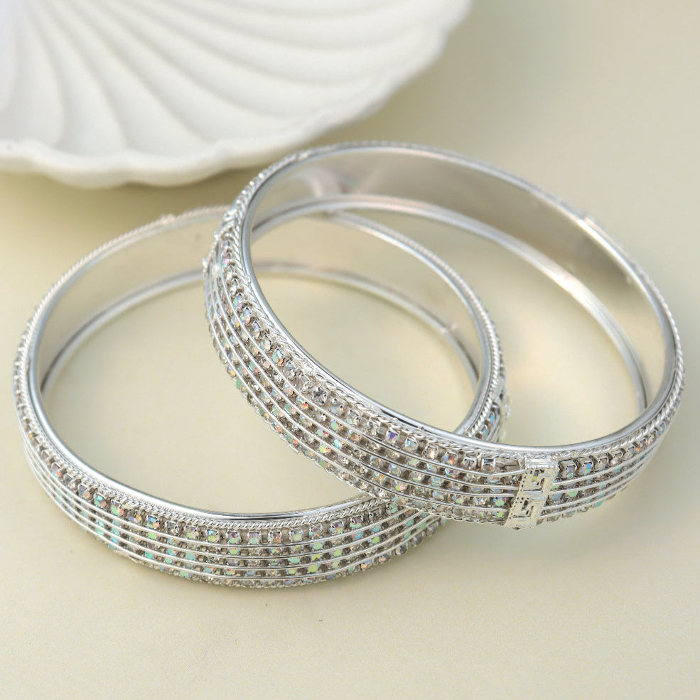 Elegant silver-toned bangles adorned with shimmering American diamonds, featuring intricate detailing and a secure clasp. Displayed against a soft pastel background with a decorative shell tray, enhancing their luxurious appeal.