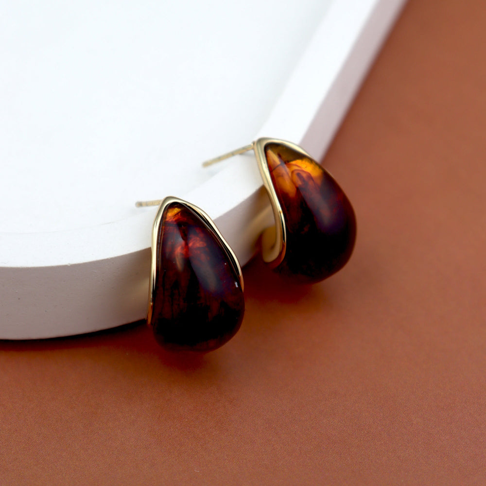 Elegant teardrop-shaped amber stud earrings with a glossy, deep brown gemstone encased in a sleek golden frame, displayed on a white tray with a warm brown background. Perfect for a classic and sophisticated look. Ideal for women’s fashion and statement jewellery collections.