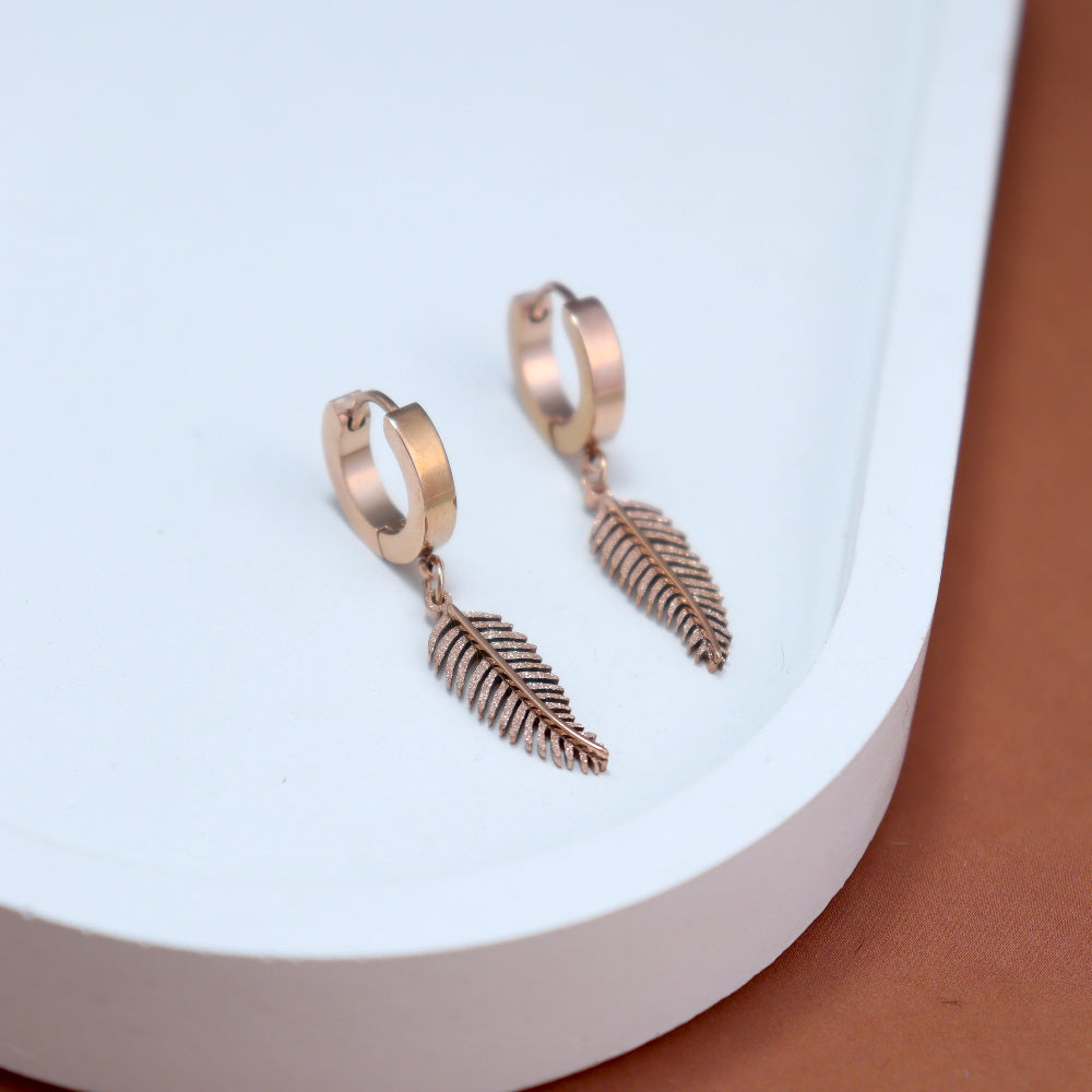 A pair of rose gold hoop earrings with delicate fern leaf-shaped dangles, displayed on a white tray with a light blue background. The earrings have a polished finish, with intricate detailing on the leaf charms.