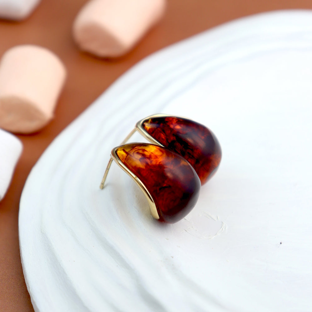 Elegant teardrop-shaped amber stud earrings with a glossy, deep brown gemstone encased in a sleek golden frame, displayed on a white tray with a warm brown background. Perfect for a classic and sophisticated look. Ideal for women’s fashion and statement jewellery collections.