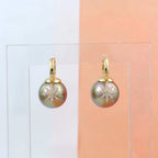 Gold earrings with greenish pearls on a clear stand against a gradient background