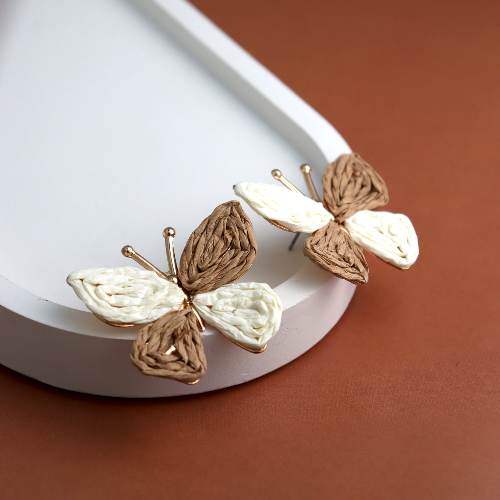 Elegant handcrafted butterfly earrings with a woven texture in neutral brown and white tones, set on a gold-tone frame. Displayed on a white ceramic tray against a warm brown background, highlighting their intricate design and artisanal charm.